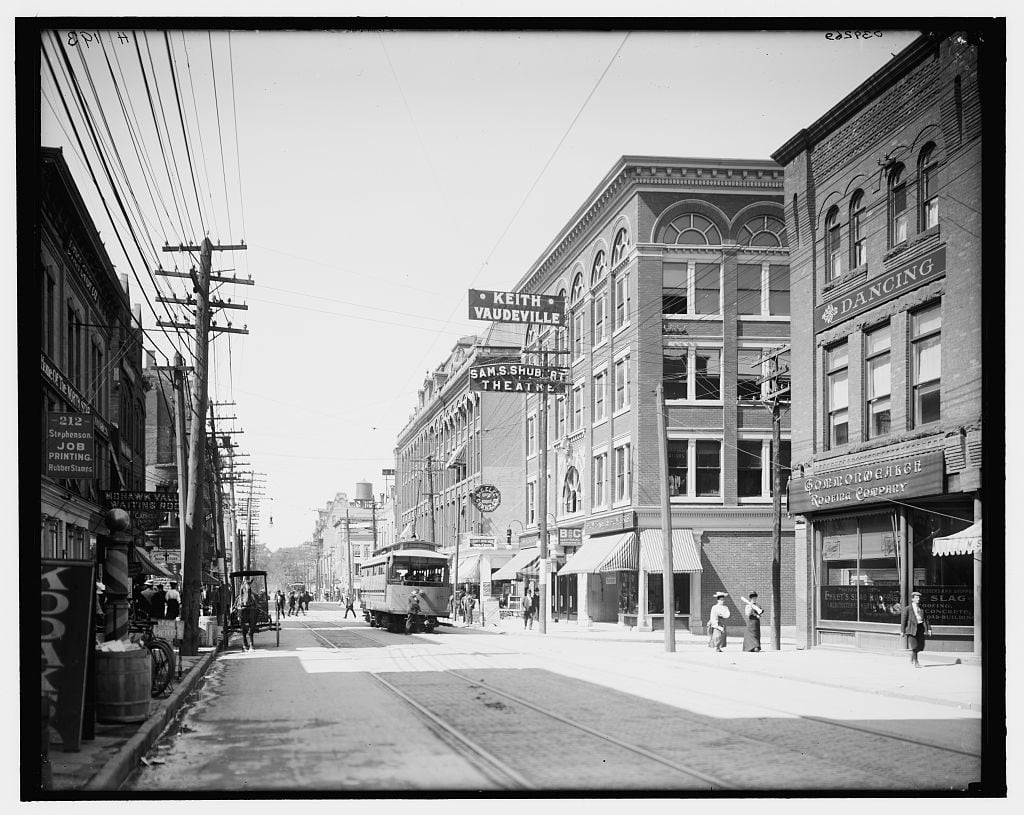 Bleecker Street in Utica in the early 1900s: storefronts with awnings, a trolley in the street, shoppers on the sidewalks, and a Keith Vaudeville sign above the buildings.