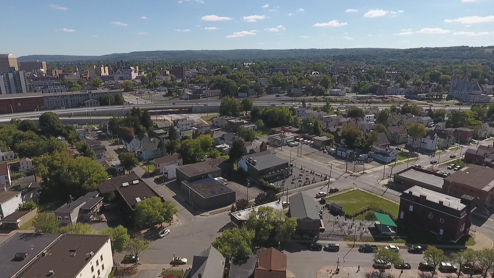 Aerial view over West Utica: streets of houses and small commercial blocks in the foreground, the elevated arterial crossing the middle distance, downtown Utica's office towers at left, and the wooded hills of the valley along the horizon.