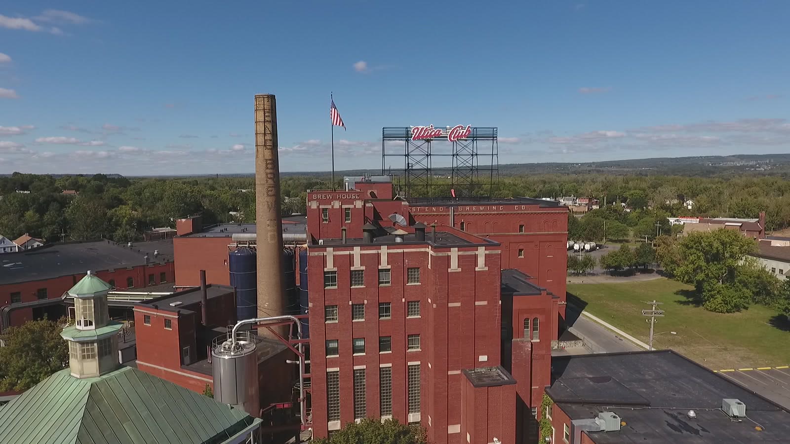 Aerial view of the F.X. Matt Brewery in West Utica: a red-brick brew house with WEST END BREWING CO. lettering along the roofline, a tall chimney beside it, and the illuminated Utica Club sign on a steel frame above the roof.