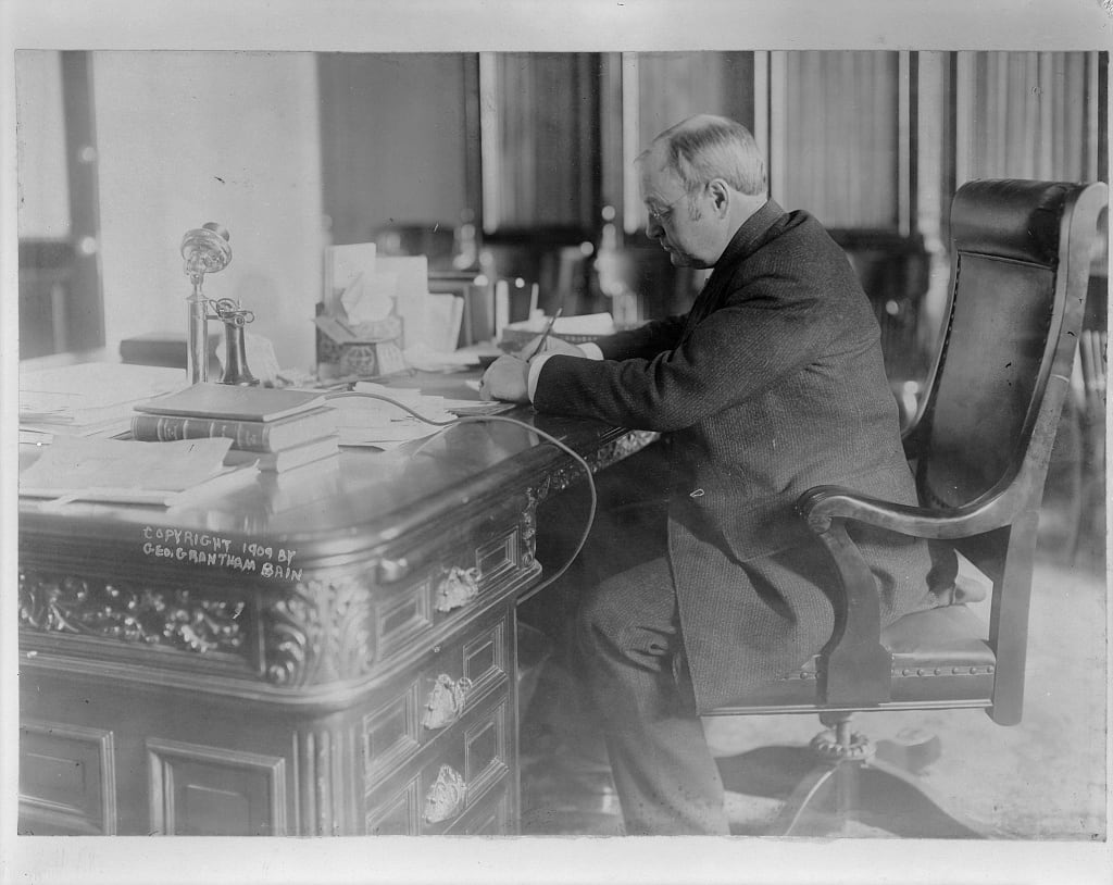 James Schoolcraft Sherman in profile at a large carved desk, writing, a candlestick telephone and stacked books beside him.