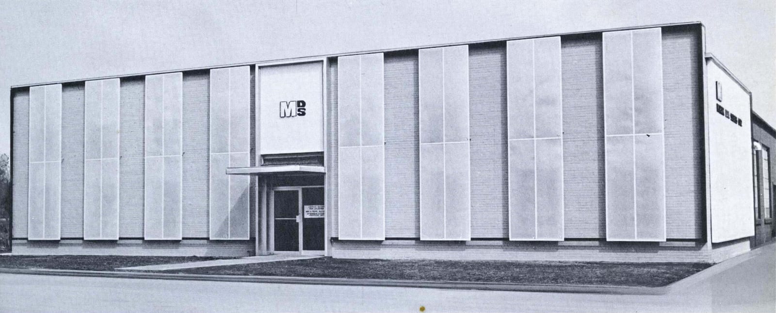 A long, flat-roofed building faced with tall pale panels, the MDS logo above its glass entrance doors and a small canopy over the door.