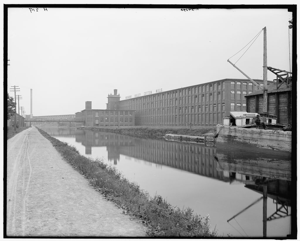 A long brick mill building labeled Mohawk Valley Cotton Mills reflected in the still water of the Erie Canal, with a dirt towpath in the foreground and a wooden barge tied up at right.