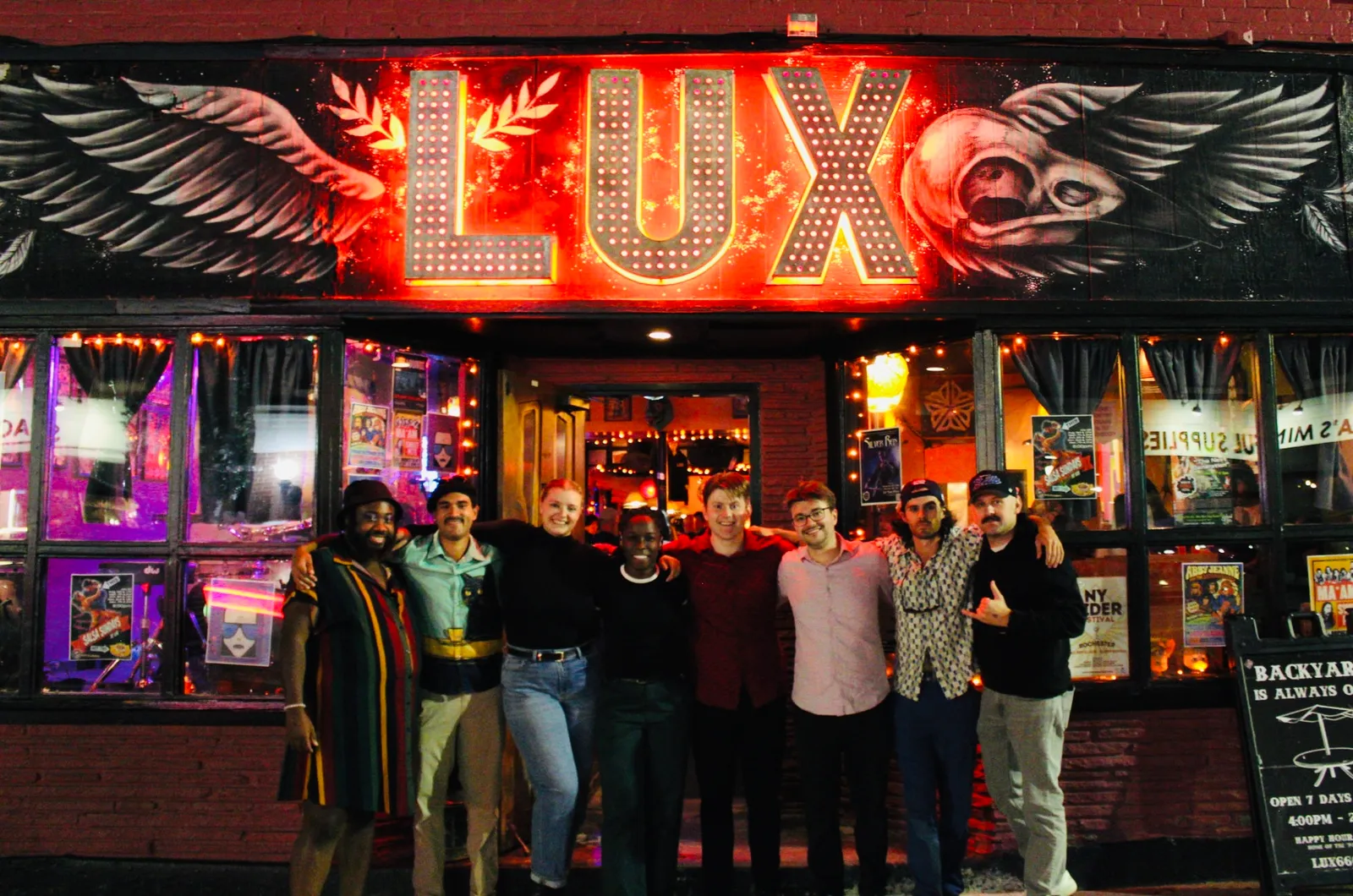 Stunt Double's eight members standing arm in arm under a lit marquee sign at night