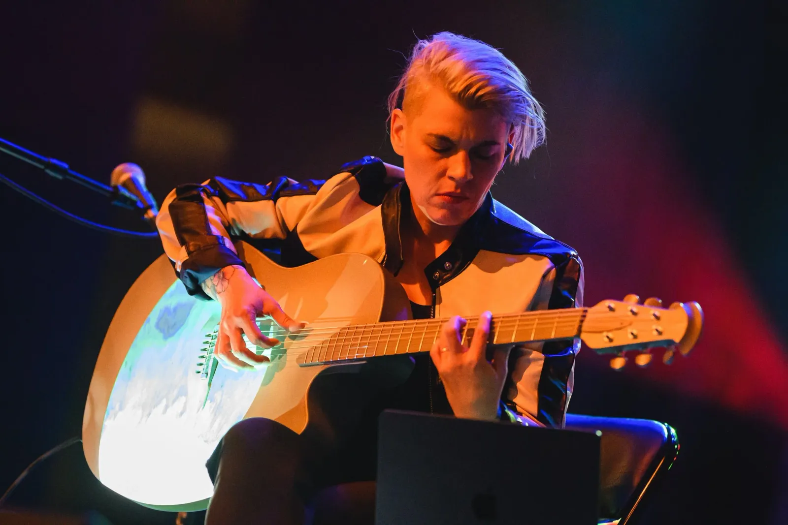 Kaki King playing an acoustic guitar on stage under red and blue light