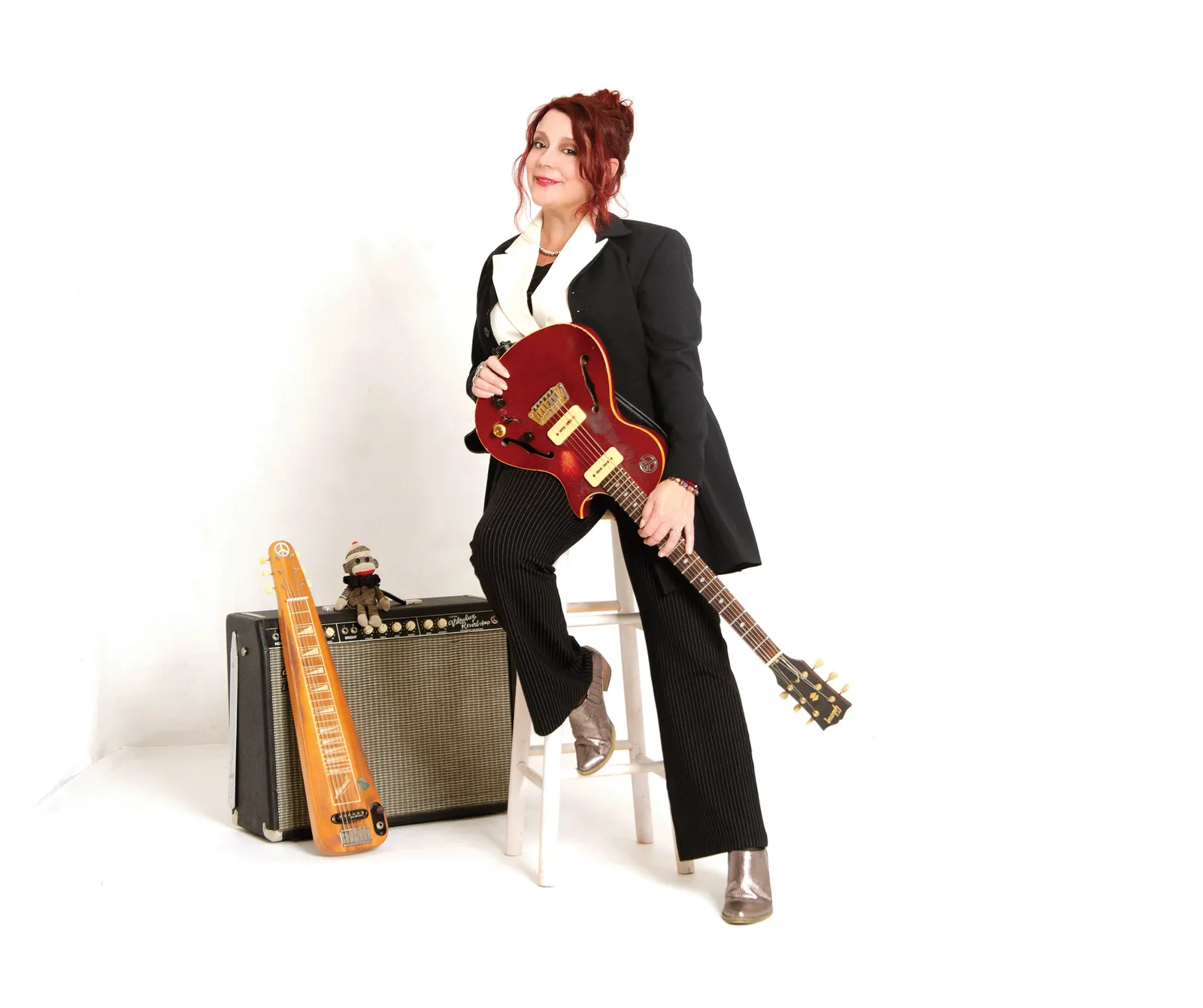 Carolyn Wonderland in a black suit on a stool with a red electric guitar, an amp and a lap steel beside her, against a white backdrop