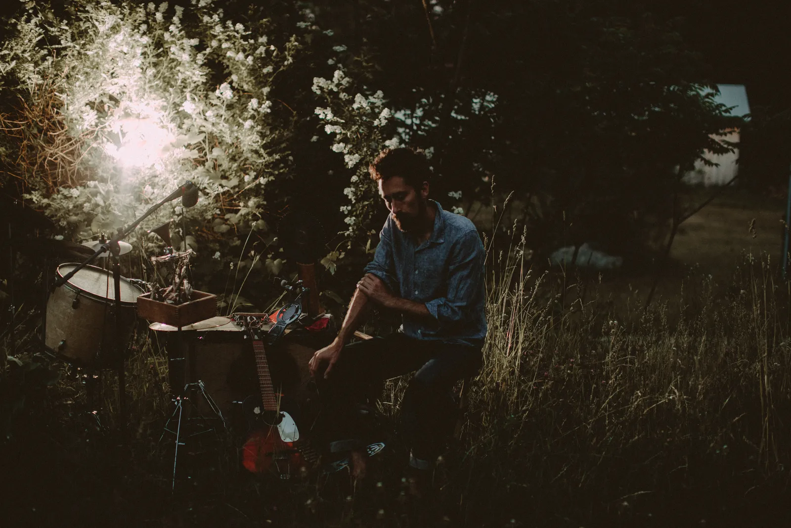 Matt Lorenz of The Suitcase Junket sitting in tall grass at dusk beside his drum, guitar and microphone