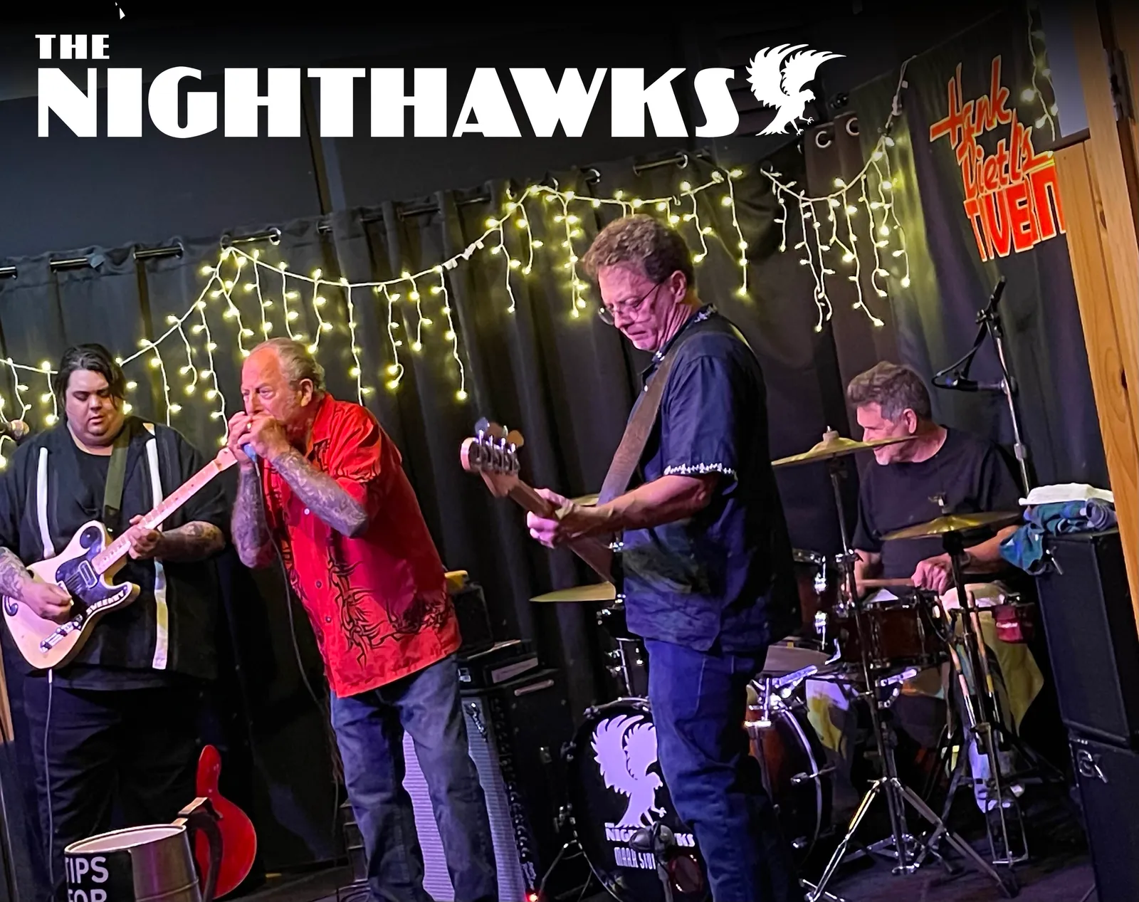 The Nighthawks playing on a small stage under string lights, harmonica, two guitars and drums, with the band's name across the top
