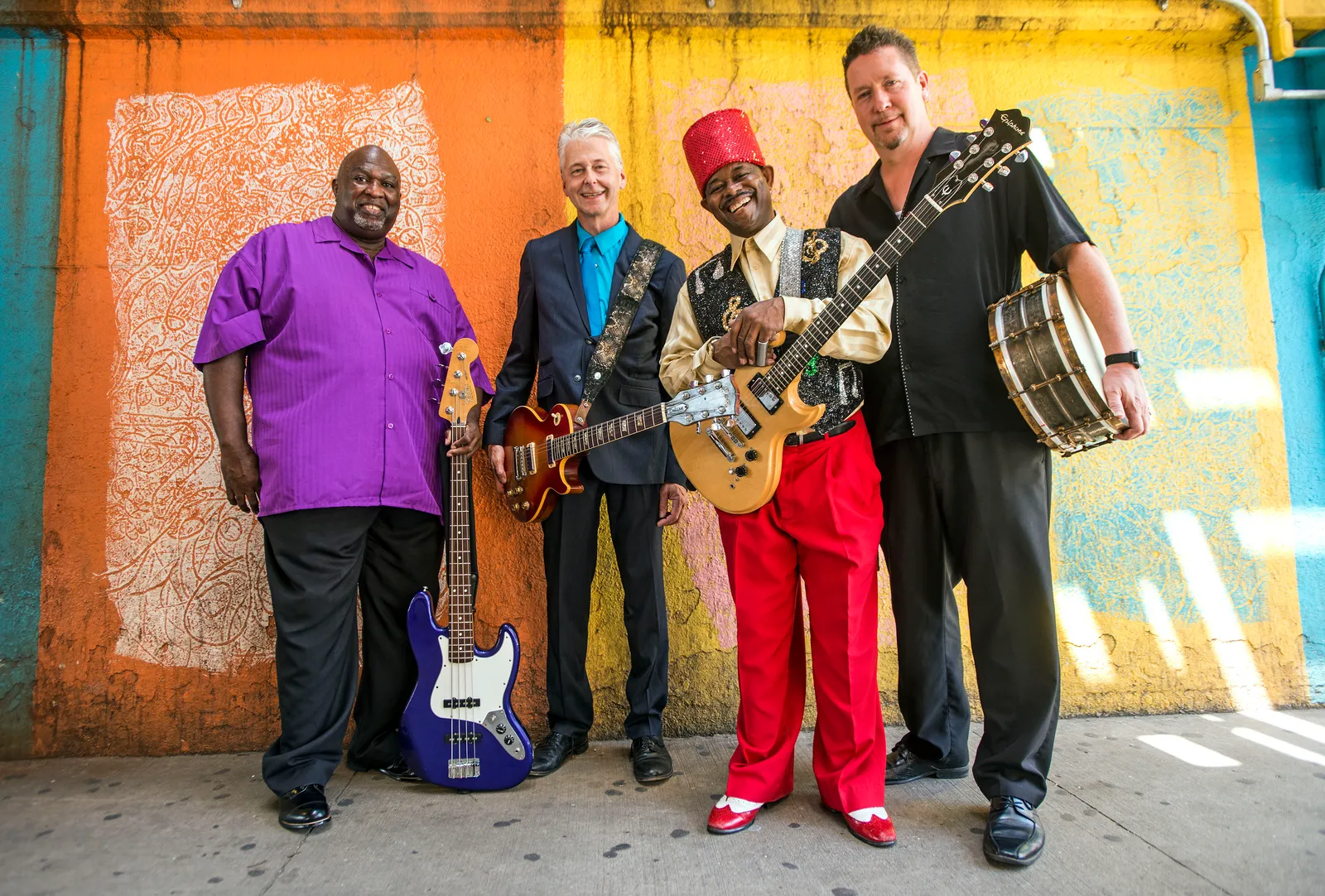 Lil' Ed & The Blues Imperials, four men with their instruments, in front of a painted orange, yellow and blue wall