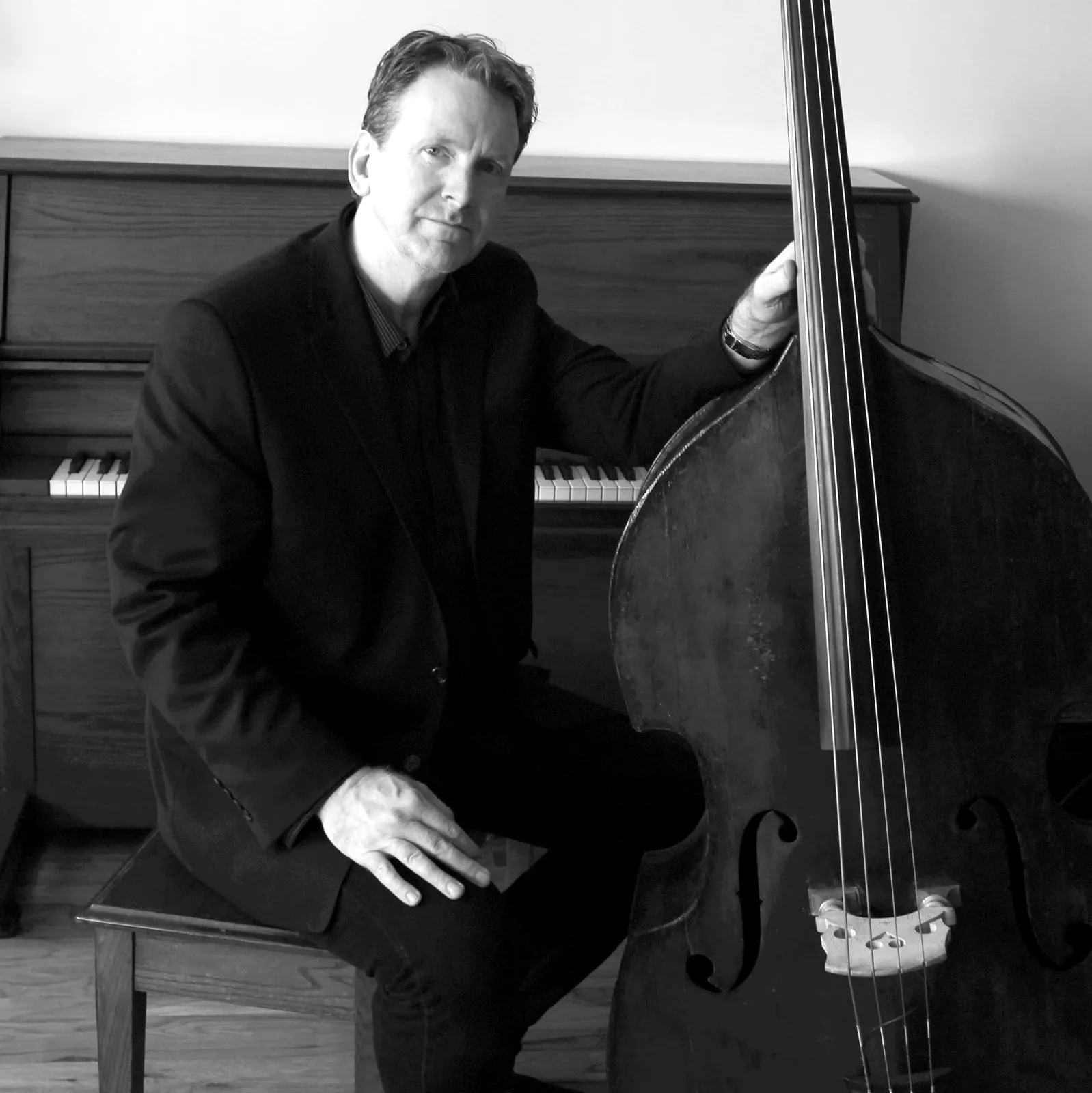 Tom Kennedy seated with an upright bass beside a piano, in black and white