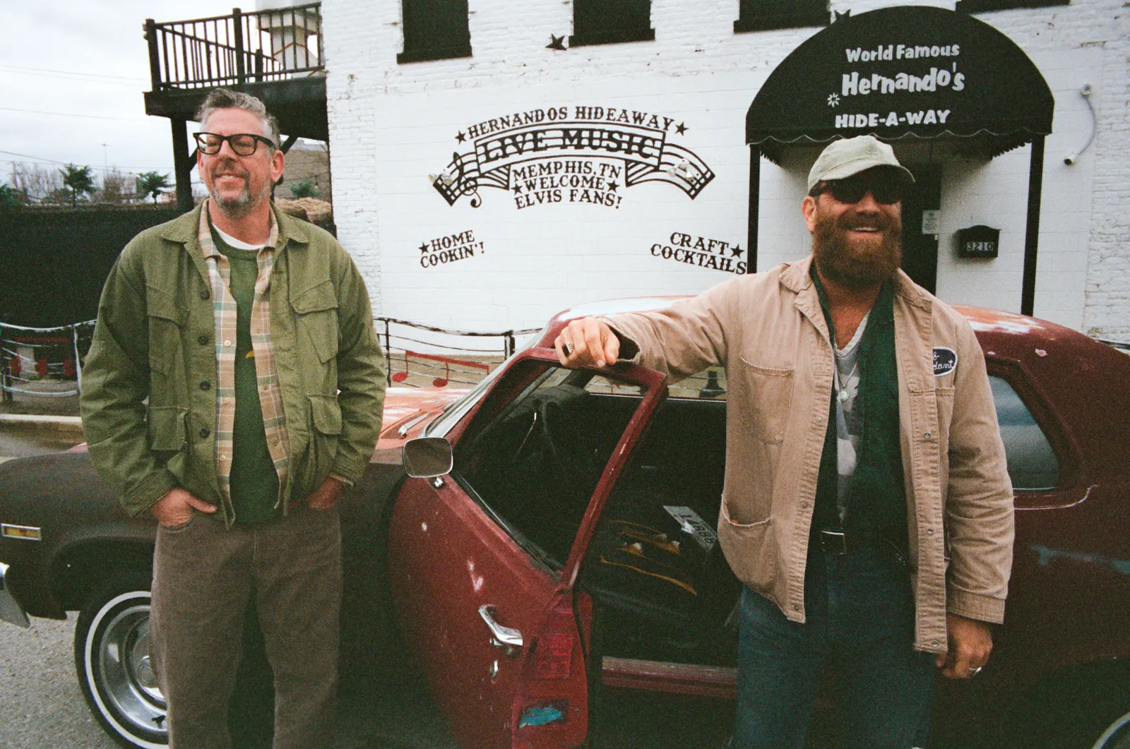 The two members of The Black Keys beside a red car in front of a white building painted with a Hernando's Hide-A-Way live music sign