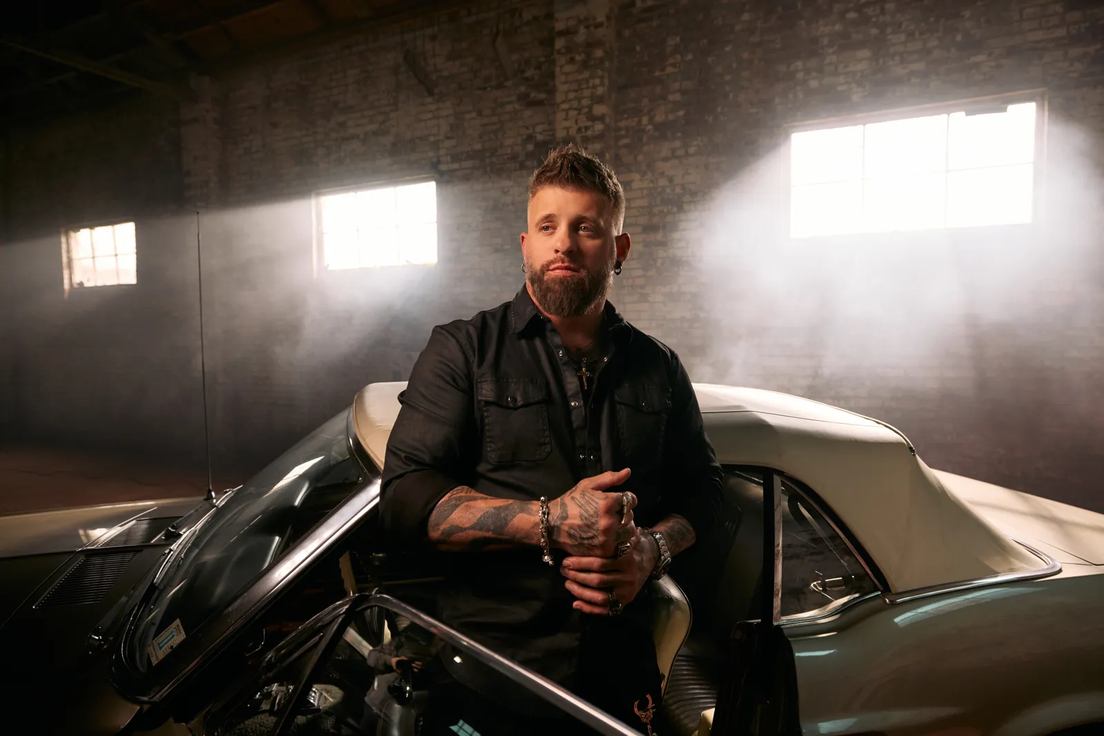Brantley Gilbert in a black shirt leaning on the door of a white convertible inside a brick warehouse, light coming through the windows