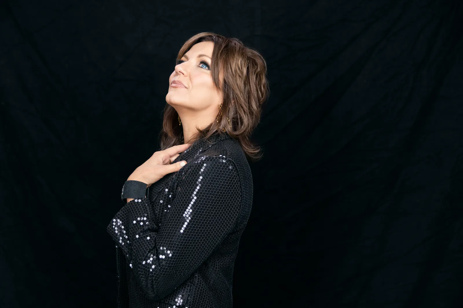 Martina McBride in a black sequined jacket against a black backdrop, looking upward with a hand at her collar