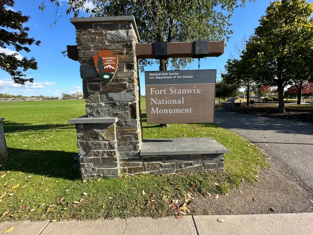 The Fort Stanwix National Monument entrance sign on its stone base, with the fort's lawn and trees behind it
