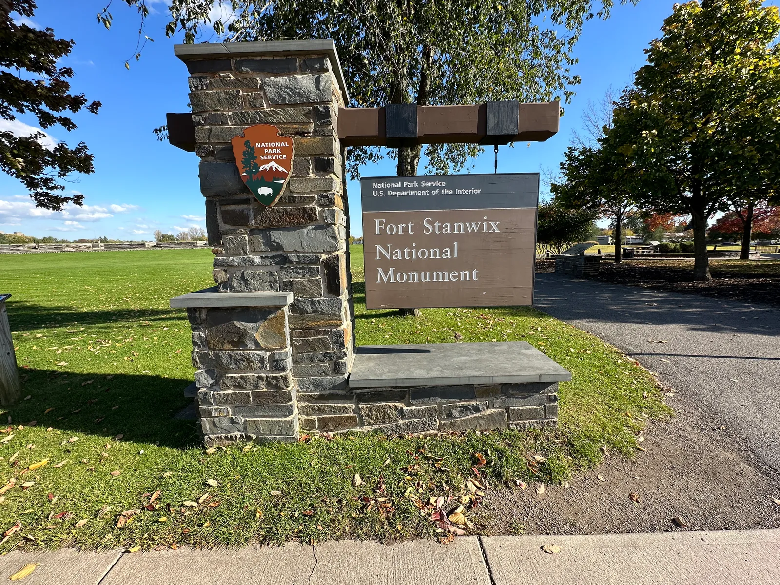The Fort Stanwix National Monument entrance sign on its stone base, with the fort's lawn and trees behind it